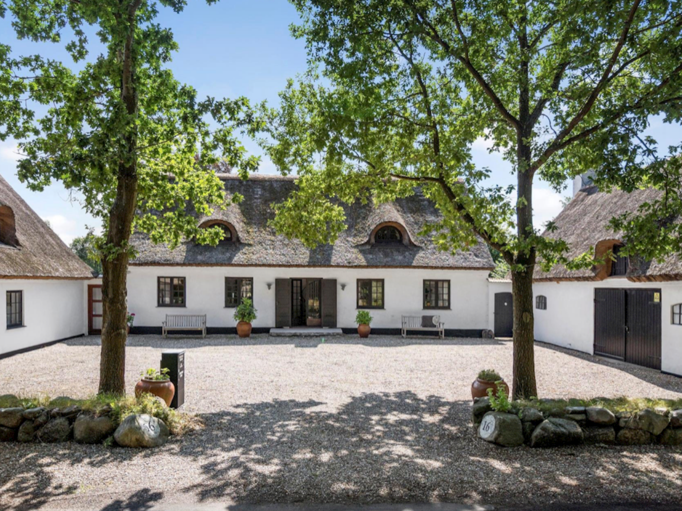 Hvid stråtækt landejendom omgivet af store træer, med gårdsplads og bænke foran indgangen, som en del af de rolige undervisningsrammer på Tobias‑Skolen